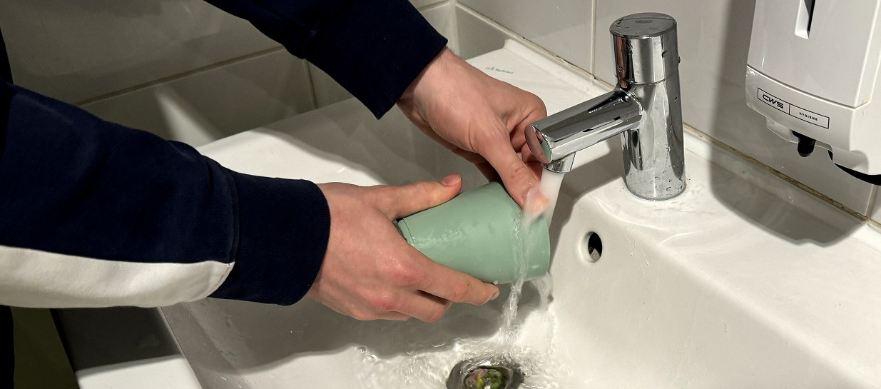 Someone rinsing a reusable cup under the tap of a washbasin, away from the pantry